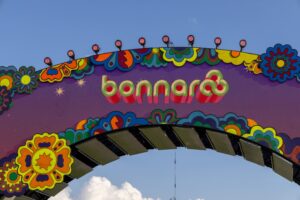 Colorful psychedelic carnival arch with ornamental floral shapes and stage lights on top against a blue sky (likely festival decor).