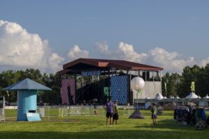 Outdoor music festival scene with a large open stage labeled Bonnaroo, blue sky and fluffy clouds, and festival-goers in the foreground.