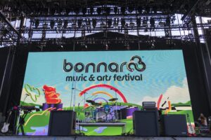 Outdoor festival stage with a large colorful LED backdrop displaying 'bonnaroo music & arts festival' and a drum kit set up on stage with instruments nearby
