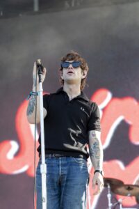 Male singer wearing a black polo and sunglasses performs on stage, gripping a microphone stand with tattoos visible on his arm.