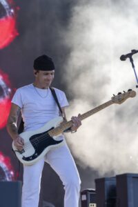 Male bassist wearing a black beanie and white outfit plays a cream Fender bass on a smoky stage. Red lights and equipment visible behind him.