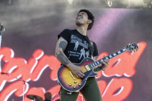 Guitarist playing an orange-to-burst sunburst Les Paul on stage with tattoos visible on his left arm, wearing a black graphic t‑shirt.