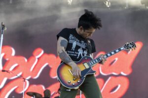 Male guitarist playing a sunburst electric guitar on stage, tattoos visible on his arm, focused on the fretboard during a live concert.