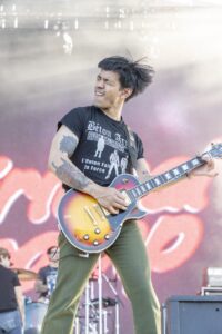 Guitarist in a black T-shirt playing a sunburst electric guitar on stage, tattoos visible on his arm.