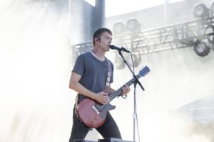 Male guitarist in a gray t‑shirt playing a red electric guitar and singing into a microphone on an outdoor stage.