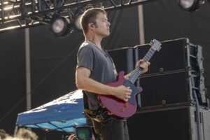 Male guitarist playing a red electric guitar on stage, wearing a gray t-shirt and in-ear monitor outdoors at a performance venue.