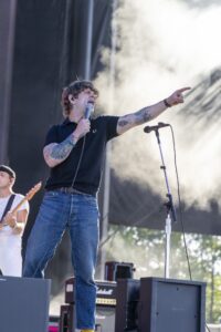 Male singer on stage wearing a black polo, singing into a handheld microphone while pointing into the crowd; visible tattoos on his arms and blue jeans, outdoor concert setting.