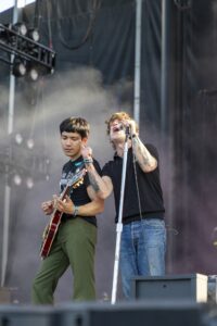 Two male musicians on stage: guitarist left, vocalist right singing into a microphone amid smoke and stage equipment.