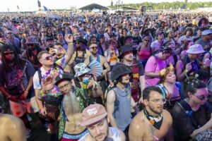 Large crowd at an outdoor music festival, people wearing colorful outfits and sunglasses, cheering and waving hands.