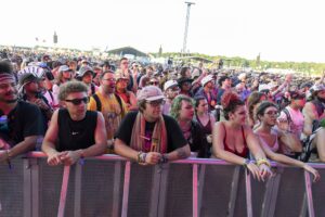 Large crowd of festival-goers behind a barrier, many wearing sunglasses and colorful outfits, with a distant stage and equipment in view.