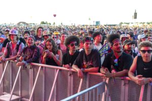 Front-row crowd at an outdoor concert behind a metal barrier, people wearing hats, sunglasses, and colorful outfits.
