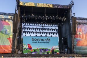 Outdoor music festival stage with colorful banners and a large LED screen reading 'bonnaroo music & arts festival' and a band performing below