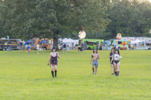 Group of festival-goers walking across a grassy field with colorful food trucks and tents in the background.