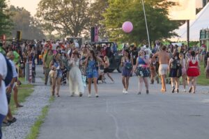 Crowd of festival-goers walking along a sunny street, many in colorful outfits, with a pink balloon floating above and trees in the background.