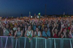 Large crowd at a nighttime outdoor concert, hands raised behind a barrier fencing off the stage area.
