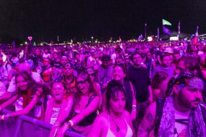 Large crowd at a nighttime festival, purple lighting, leaning on a barrier and cheering.