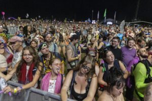A crowded festival crowd behind barrier at night, people dancing and taking photos in colorful outfits.