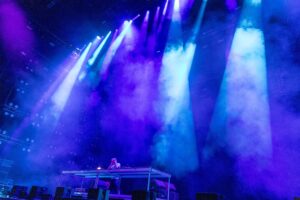 DJ on an elevated booth performing on a concert stage with blue and purple lights and fog.