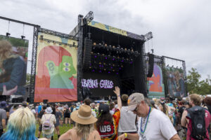 Outdoor music festival crowd facing a large stage with colorful banners and lighting rigs under a cloudy sky