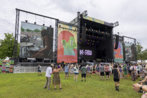 Outdoor music festival with a large stage, colorful banners, and a crowd on the grass watching performers on big screens