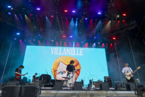 Concert stage with musicians performing; a bright blue screen shows a stylized woman’s face and the word 'VILLANELLE' above.