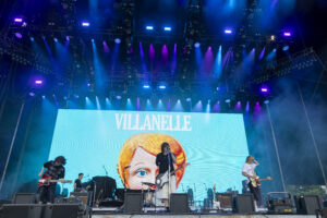 Band performs on an outdoor stage with a large LED screen showing a cartoon portrait and the text 'VILLANELLE'; blue and purple stage lights wash the scene.