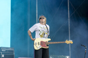 Guitarist wearing sunglasses and a white graphic tee plays a cream bass on a blue stage backdrop.