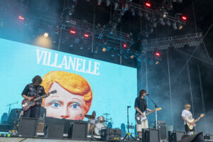 Band performs on a large outdoor stage; a bright blue screen shows a cartoon woman's face and the word 'VILLANELLE' while guitarists play in front of it.