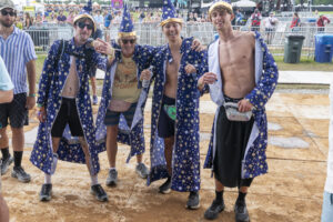 Group of men in starry blue robes posing arm-in-arm at a festival, smiling under sunny skies.