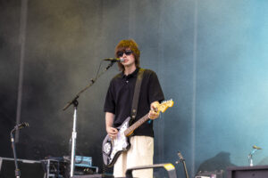 Male guitarist on stage wearing a black shirt and sunglasses, singing into a microphone with a teal backdrop behind him.