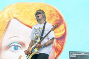 Male guitarist wearing sunglasses and a white graphic T-shirt plays a yellow bass on stage, with a large colorful mural behind him.