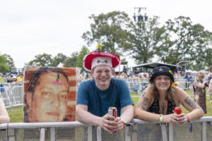 Smiling man in a blue shirt wearing a red hat with a flower, holding a can at a festival barrier beside a tattooed woman.