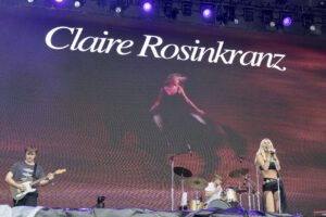 Concert stage with musicians performing; large screen behind reads 'Claire Rosinkranz'; female singer at right, guitarist on left under purple lights.
