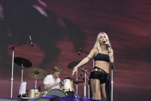 Female singer with blonde hair performing on stage, holding a microphone, wearing a black crop top and turquoise jewelry with a drummer in the background.