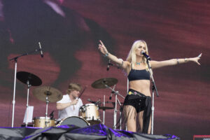 Female singer on stage with mic, arms extended, performing in a concert with a drummer in the background.