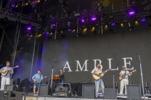 Band performing live on an outdoor stage with multiple guitarists and microphones, under purple and yellow lights, backdrop with letters behind them.