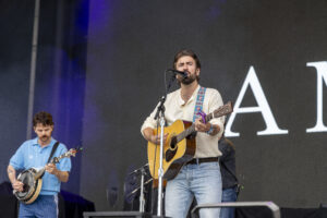 Bearded man in beige shirt sings into a microphone while playing an acoustic guitar on stage; a second musician with a banjo is visible to the left.