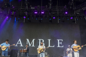 Band performs on a dim stage with multiple guitarists, microphones, and purple stage lighting in front of a large backdrop that reads AMBLE or similar text.