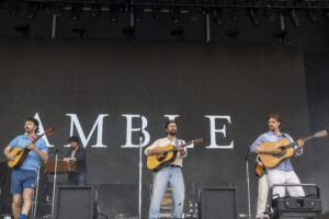 Four male musicians playing acoustic guitars on a stage with a dark backdrop and large white lettering behind them.