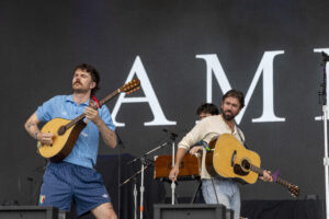 Two male musicians play acoustic guitars on a stage during a live concert, with a large dark backdrop behind them.