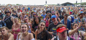Large diverse crowd at an outdoor music festival, many people wearing colorful outfits and hats