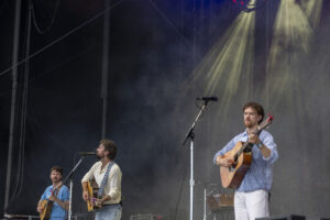 Three male musicians performing on an outdoor stage with acoustic guitars and microphones, smoke in the air behind them.