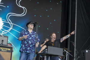 Male vocalist wearing a black hat and sunglasses performs on a stage with a blue starry backdrop and bandmates nearby.