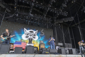 Musicians perform on a stage with a large cat-with-glasses backdrop and an American flag on the keyboard setup in front.