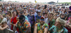 A dense, diverse crowd at an outdoor music festival near the stage, many smiling, wearing colorful clothing and sunglasses.