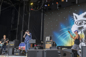 Rock band performing on an outdoor stage with a lead singer in a blue floral shirt, guitarist left, and a large cat-graphics backdrop behind them.