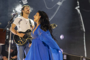 Female singer in a bright blue dress performing on stage with a guitarist nearby under stage lights.