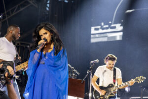 Female singer in a bright blue outfit performs into a microphone on stage, with a guitarist and bassist visible in the background.
