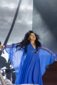 Female singer in a flowing royal blue dress performs on stage with arms outstretched and microphone in hand.