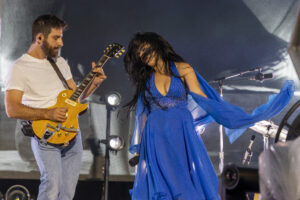 Guitarist in a white T-shirt plays a yellow Gibson on stage while a dancer in a blue dress swirls beside him.